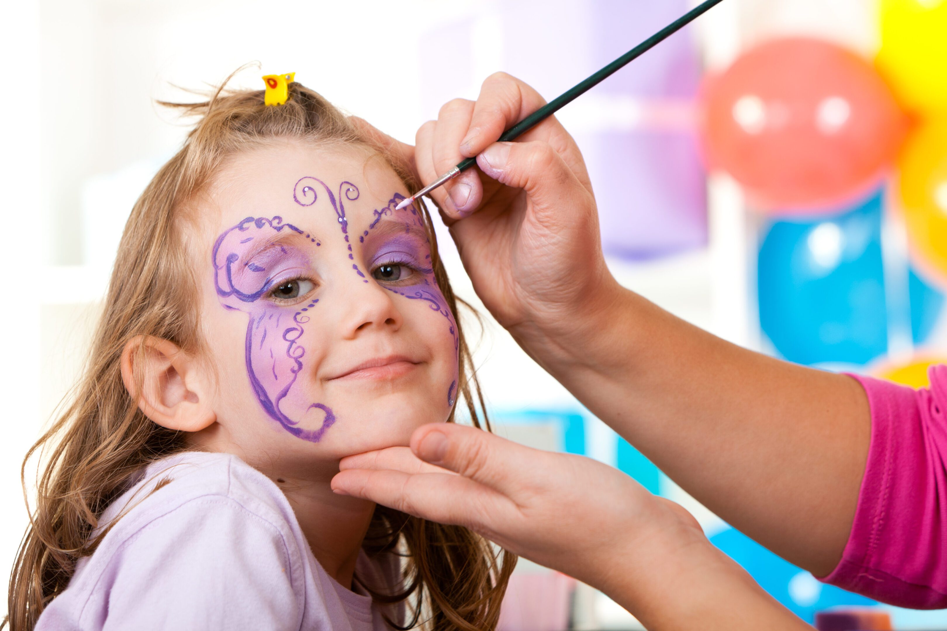 A girl getting her face painted with a purple butterfly design.