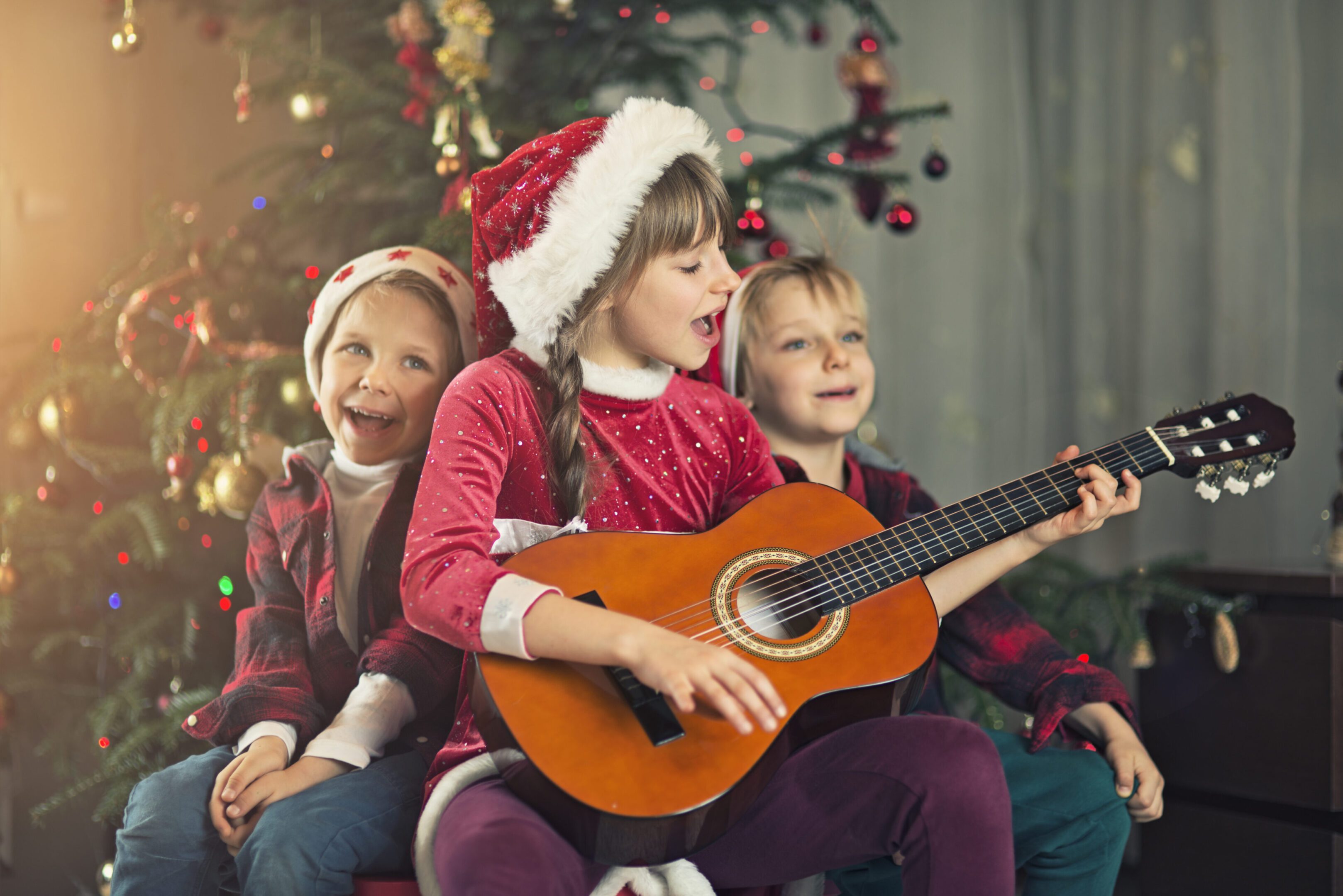 Children in Santa hats playing guitar and singing by a Christmas tree.