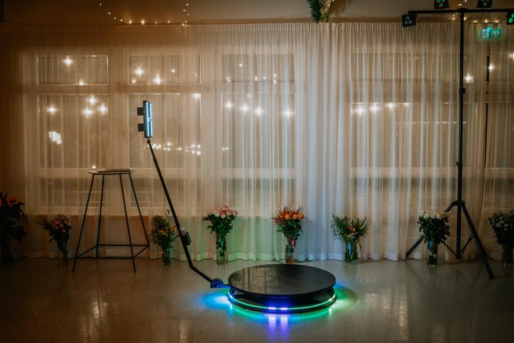 A 360-degree photo booth setup with LED lights and a rotating platform, surrounded by floral arrangements, in front of sheer curtains and string lights.