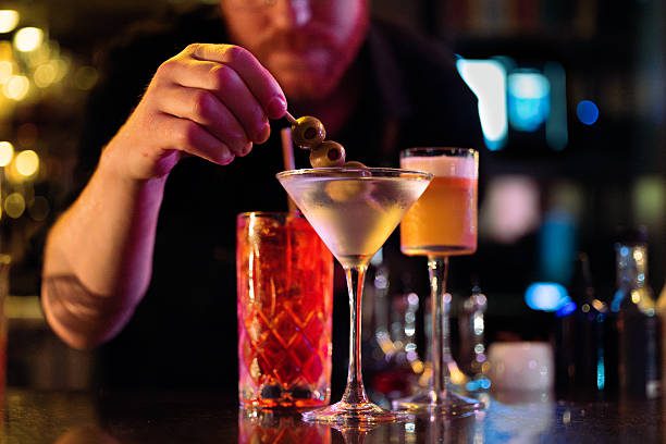 In the foreground a hand places a toothpick with three olives into a martini glass in a darkly lit bar. Behind are two other vibrant cocktails, and out of focus in the background is the rest of the bar lit in purples, yellows, and blues. The three glasses gleam on the counter.