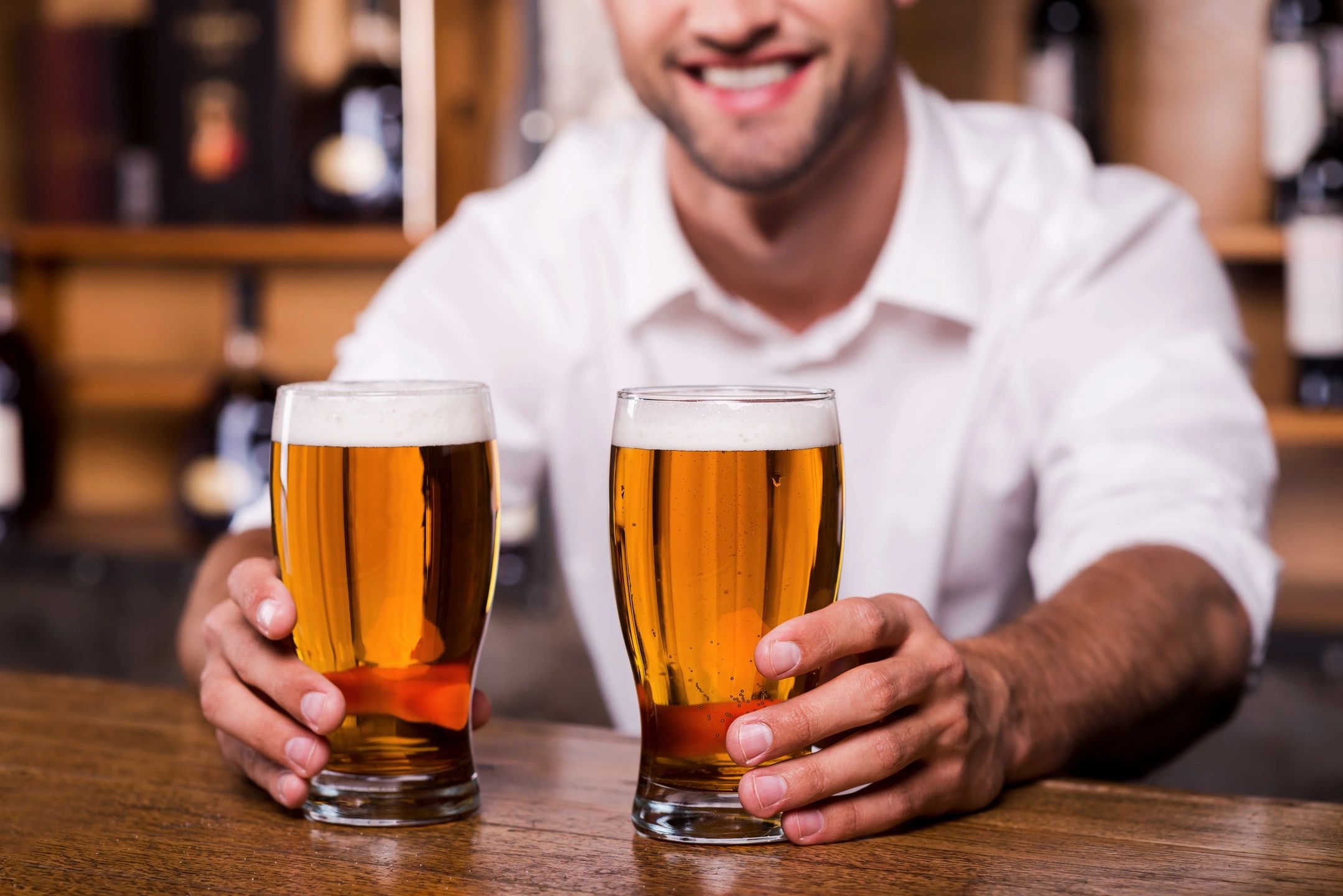 Man smiling and holding two glasses of beer at a bar.