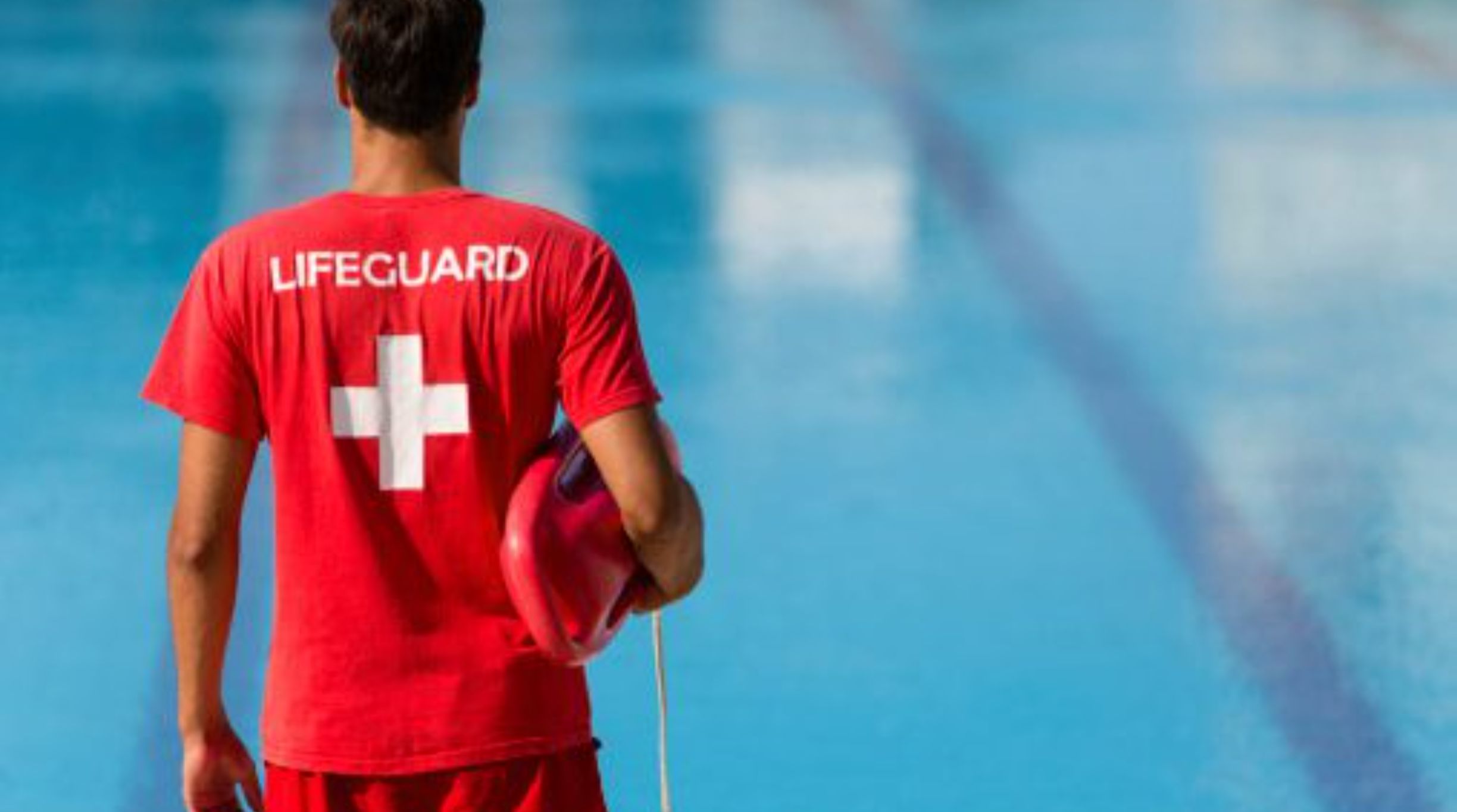 Lifeguard in red uniform holding a rescue tube by the pool.