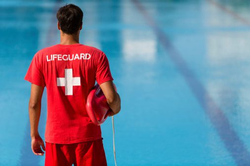 A lifeguard in a red uniform stands by a pool holding a rescue buoy.