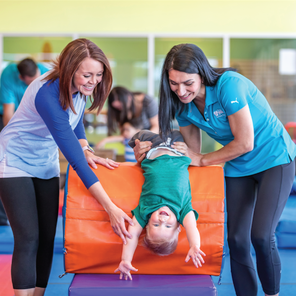 Two women help a child play on an orange padded structure indoors.